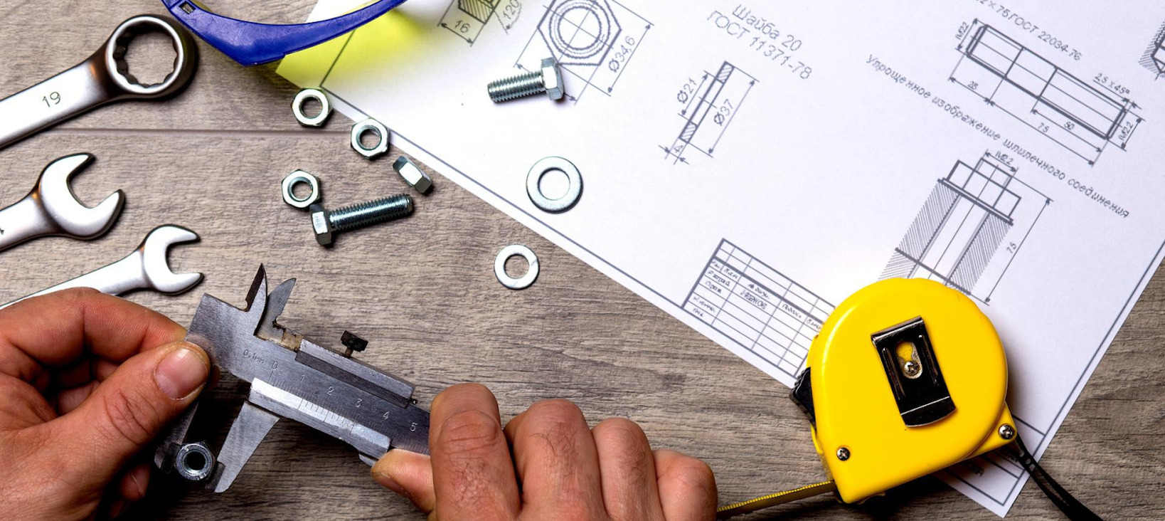 A photo of somebody measuring the size of a bolt, with several screws, spanners, and a schematic drawing of bolts in the background.