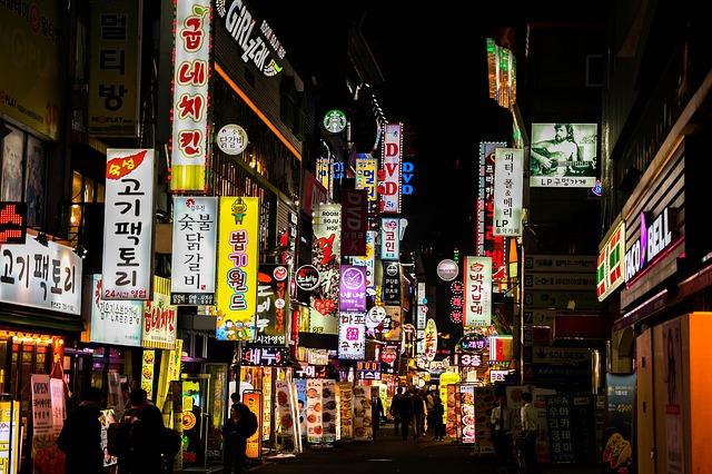 A photo of a street at night with many illuminated signs for various shops, with text in Korean.