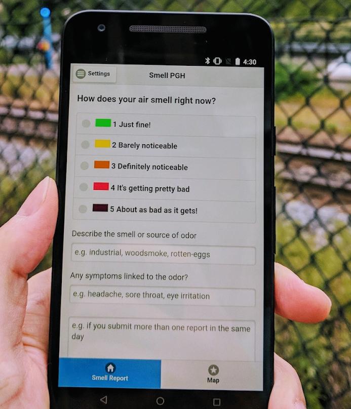 A photo of a hand holding a smart phone, which shows an app with a prompt "How does your air smell right now" with several selections from "Just fine" to "About as bad as it gets"