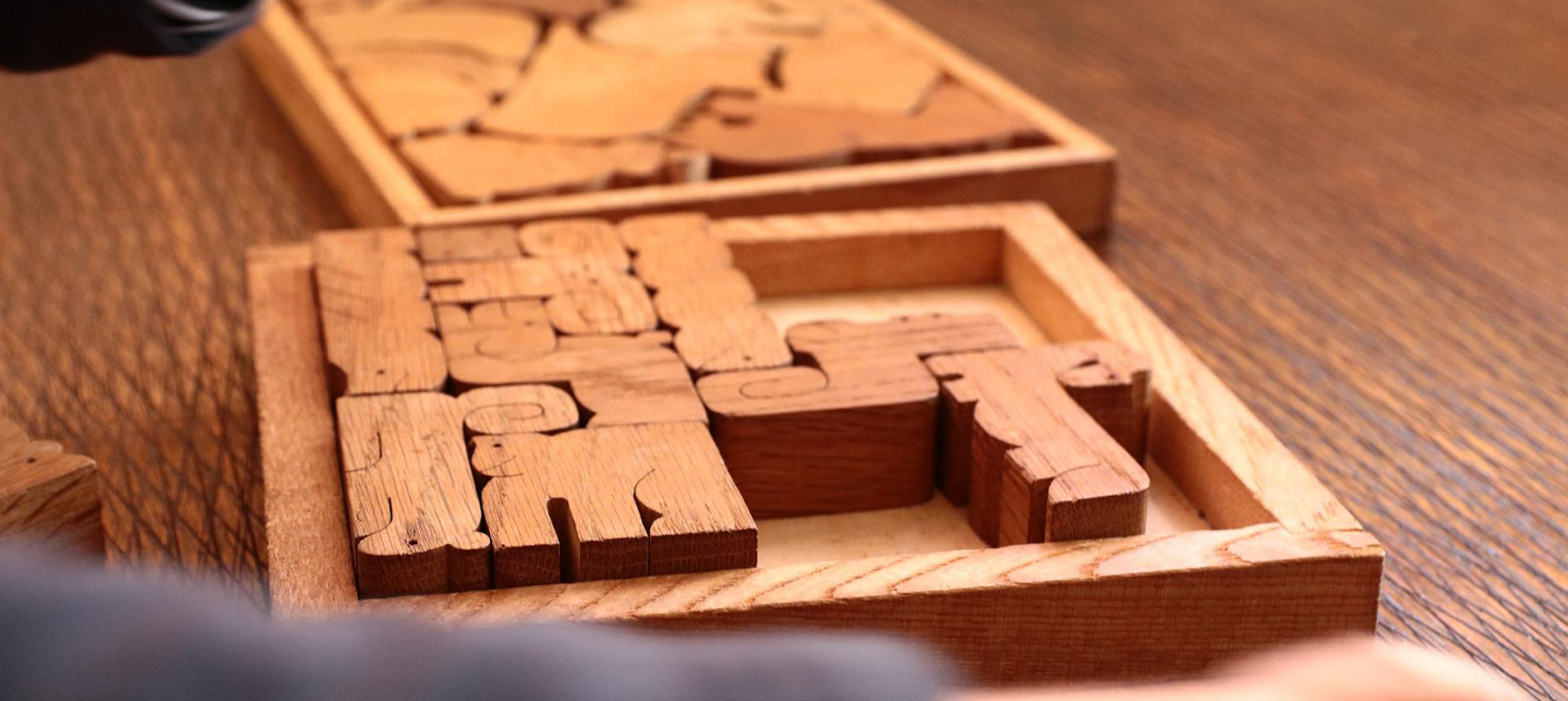 A photo of a half completed wooden puzzle with few large pieces all in the shape of animals. Hands are visible in the foreground and a completed puzzle in the background.