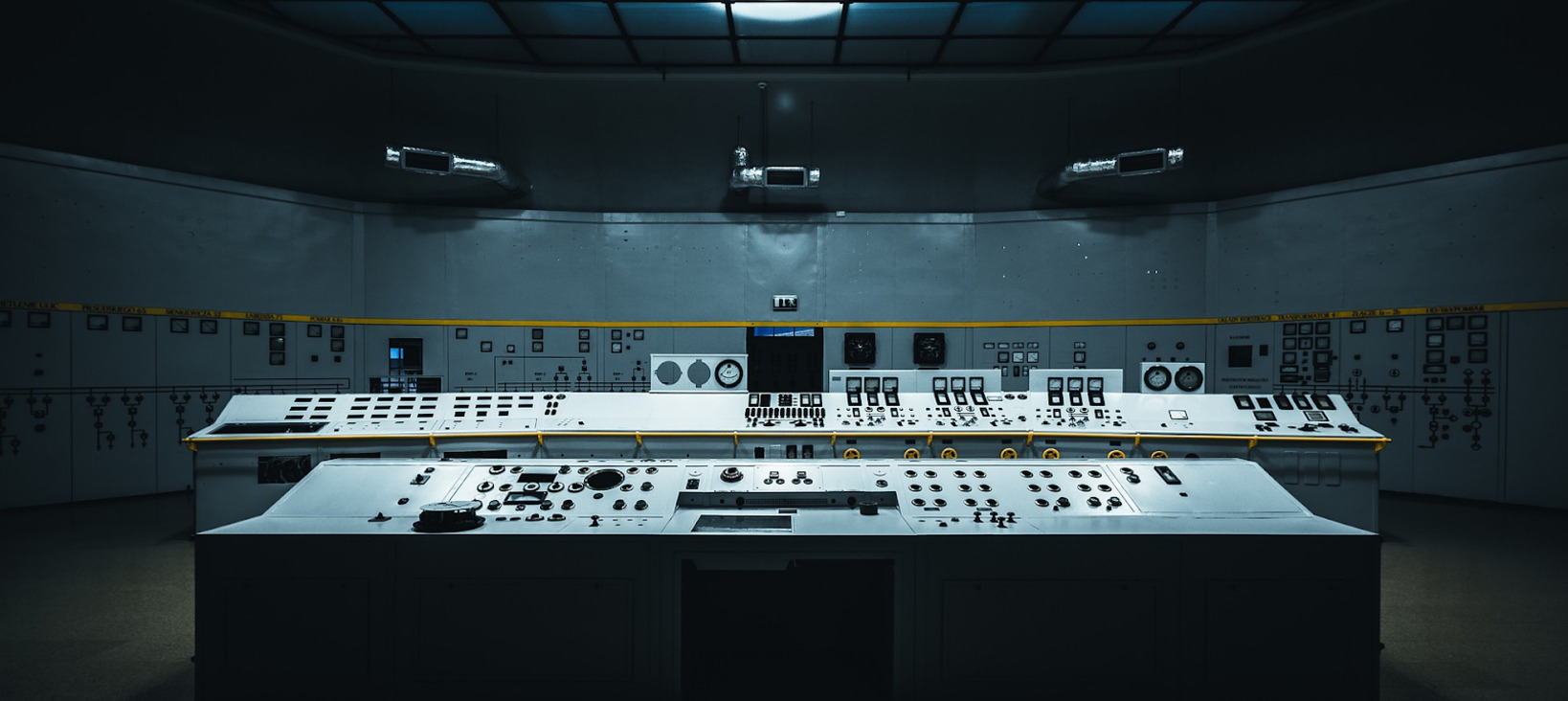 A photo of a large, windowless, dark room with a long control console with numerous dials and switches and displays. The room appears to be a control room or a space for monitoring and operating a large production system.