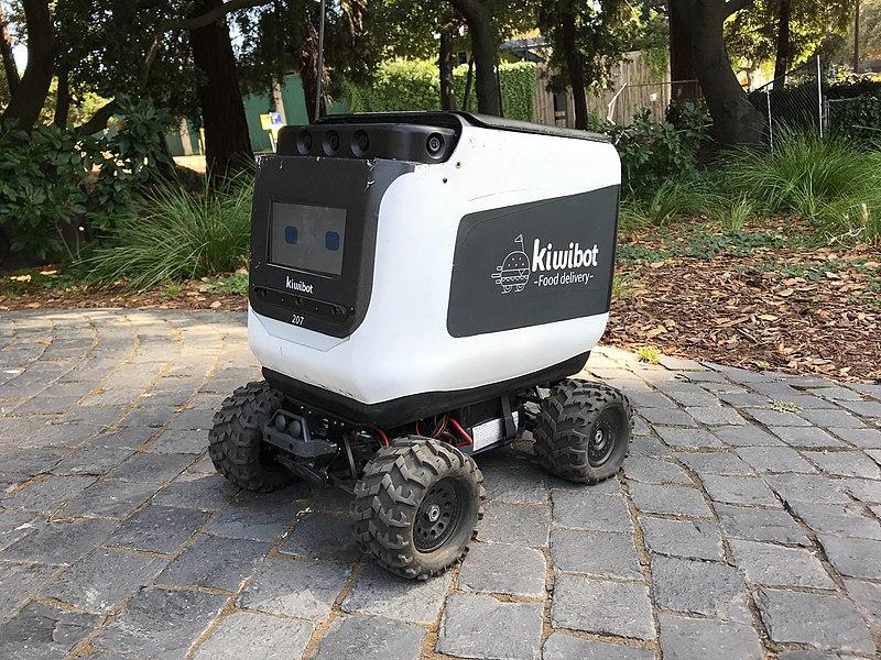 A photo of a sidewalk delivery robot labeled "kiwibot - food delivery". The robot is fairly small with four wheels and has a forward facing display showing two eyes.