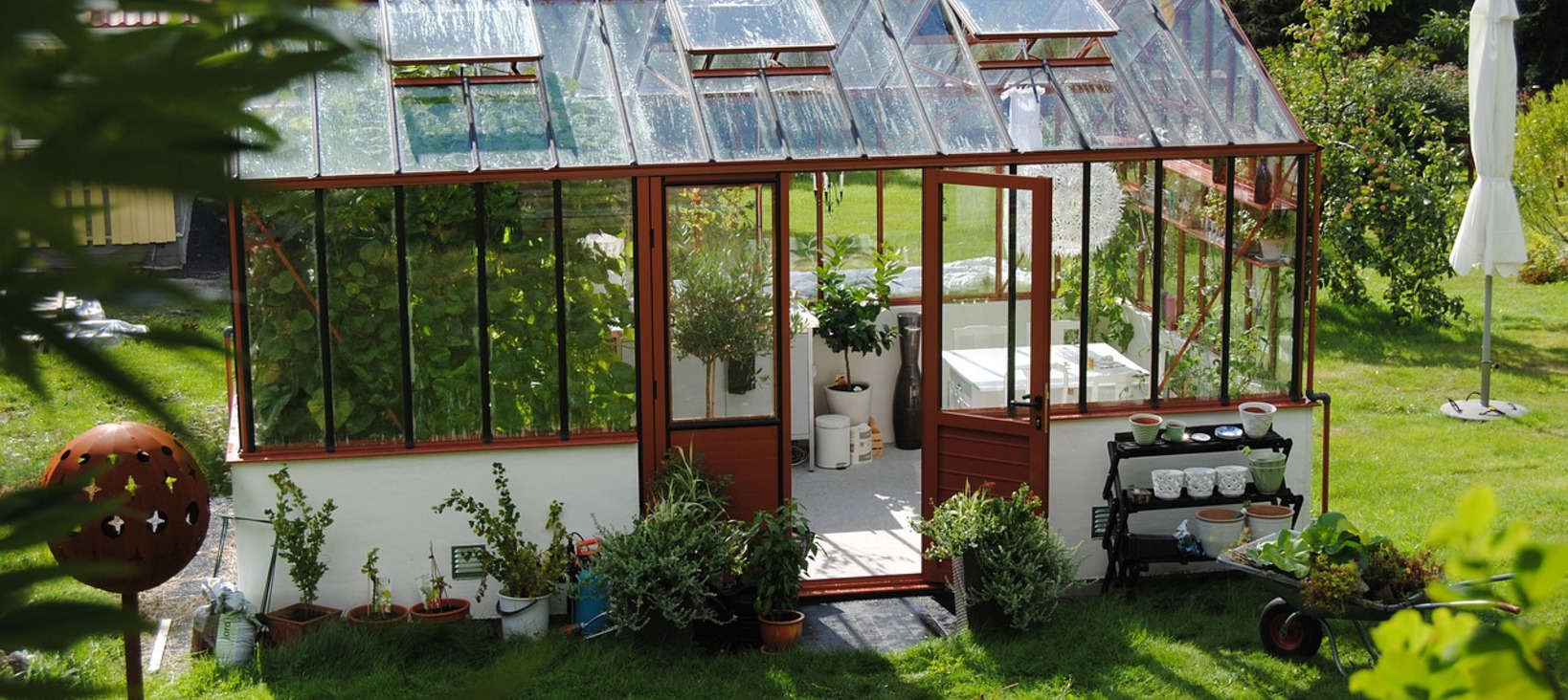 A photo of a white, glass-walled greenhouse situated in a garden. The house is surrounded by various potted plants, creating a lush and green atmosphere.