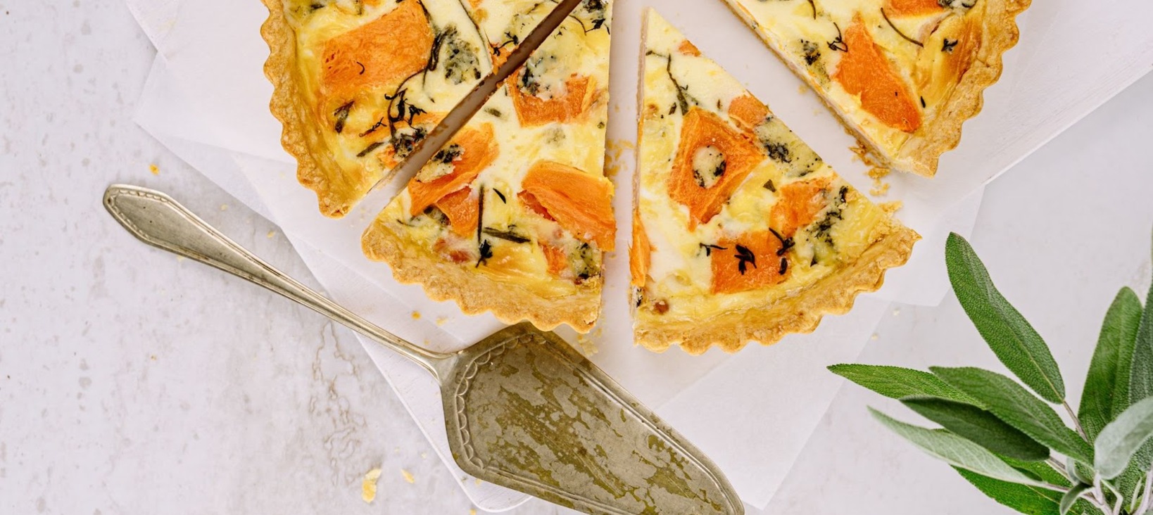 A photo of a savory pie or quiche, cut into several slices on a white counter top. Next to the pie is a serving spoon and some sage leaves.