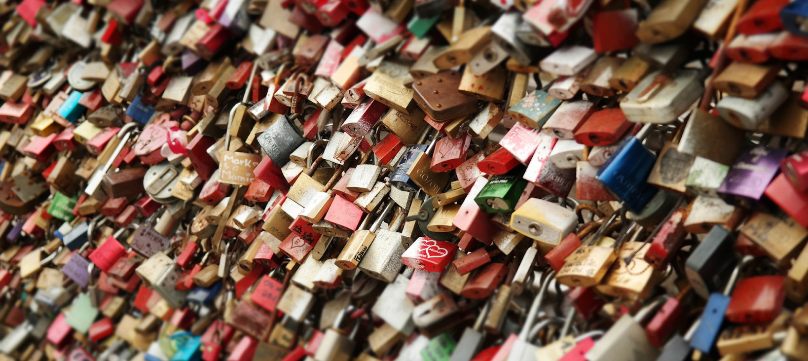 A photo of a very large number of locks attached to a structure like a fence that is not visible because it is entirely covered by the locks. Some locks have writing or heart symbols on them.
